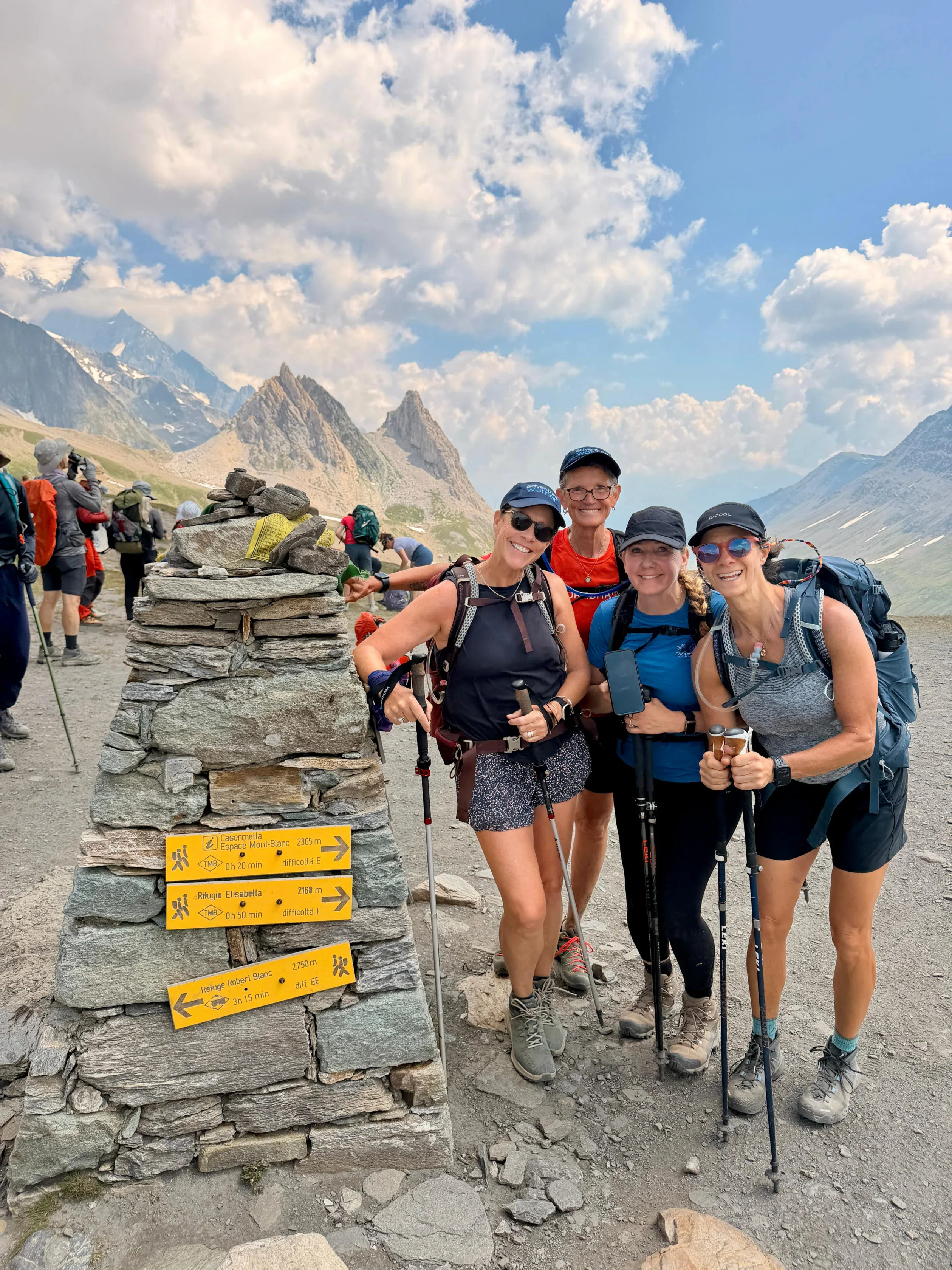 Four hikers with trekking poles stand smiling beside a stone trail marker with yellow signs in a mountainous landscape under a partly cloudy sky.