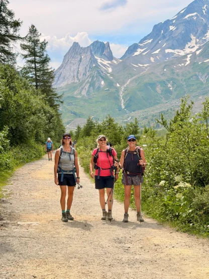 Three women with hiking gear walk on a dirt trail through green mountains, with tall peaks and trees in the background. A fourth hiker walks further behind them.