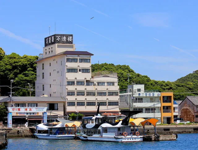 A waterfront scene shows several boats docked by a multi-story building with Japanese signage, with trees and blue sky in the background.