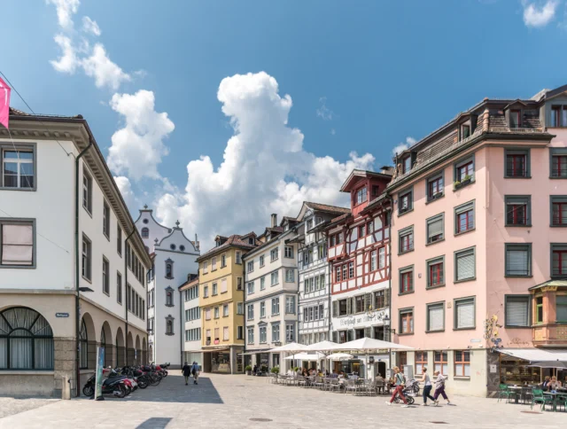 A sunny European square with colorful multi-story buildings, outdoor café seating, and people walking on a cobblestone street under a partly cloudy sky.