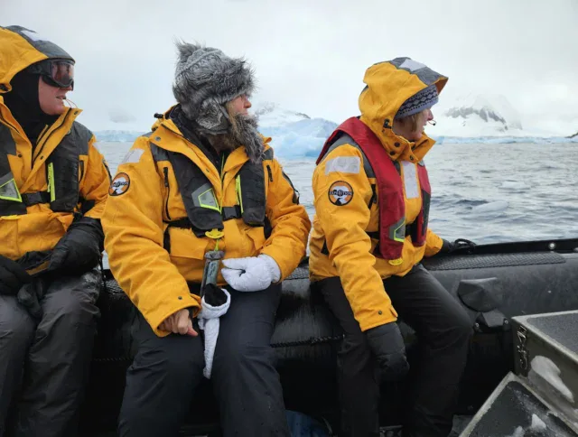 Three people in yellow parkas sit on a small inflatable boat in cold, icy conditions with snow-covered mountains in the background.