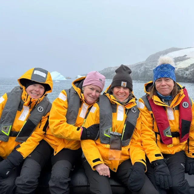 Four people in yellow jackets and life vests sit on a boat in a cold, icy environment with water, rocks, and snow in the background.