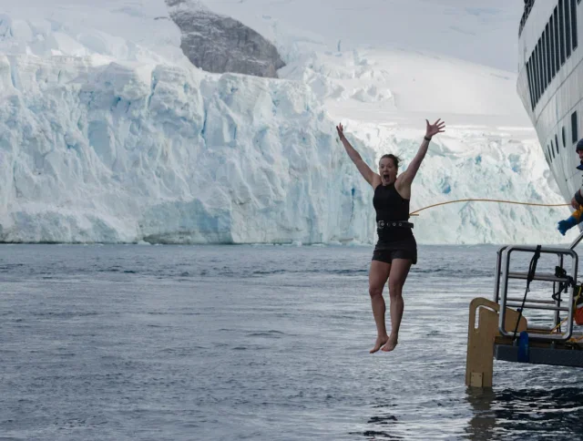 A woman in swimwear jumps off a ship into icy water near a glacier, while three people watch from the vessel.