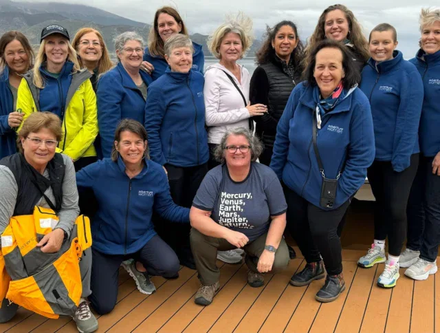 A group of fifteen women stand and kneel on the deck of a boat with water and mountains in the background, smiling at the camera.