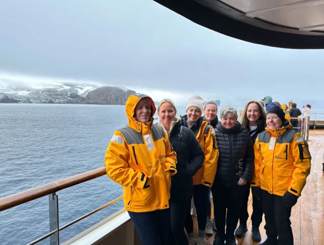 A group of people in yellow jackets stands on a ship's deck with a snowy landscape and ocean in the background.