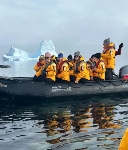 A group of people wearing yellow jackets sit in an inflatable boat on calm water with icebergs in the background.