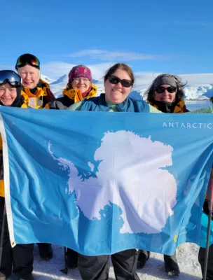 A group of people in yellow jackets hold an Antarctica flag on a snowy landscape with mountains in the background.