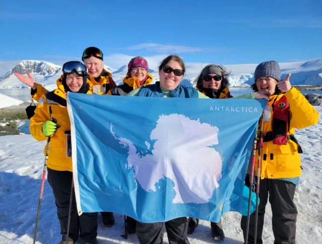 A group of people in yellow jackets hold an Antarctica flag on a snowy landscape with mountains in the background.