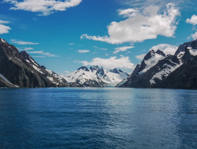 A view of snow-capped mountains and calm blue water under a partly cloudy sky.