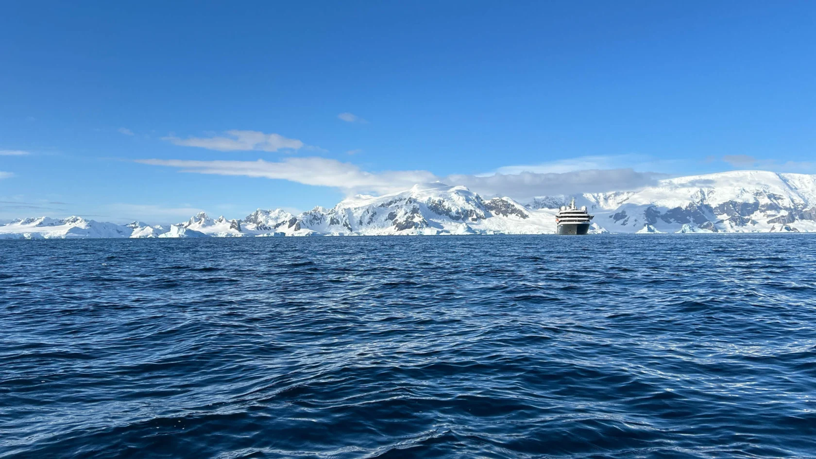 A ship sails in the ocean with a backdrop of snowy mountains and icebergs under a clear blue sky.