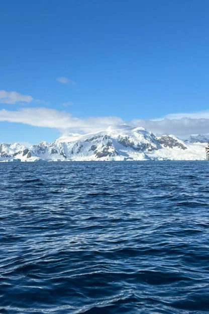 A ship sails in the ocean with a backdrop of snowy mountains and icebergs under a clear blue sky.