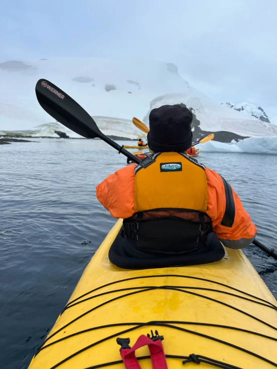 Person in a yellow kayak paddles through icy waters near snowy mountains.