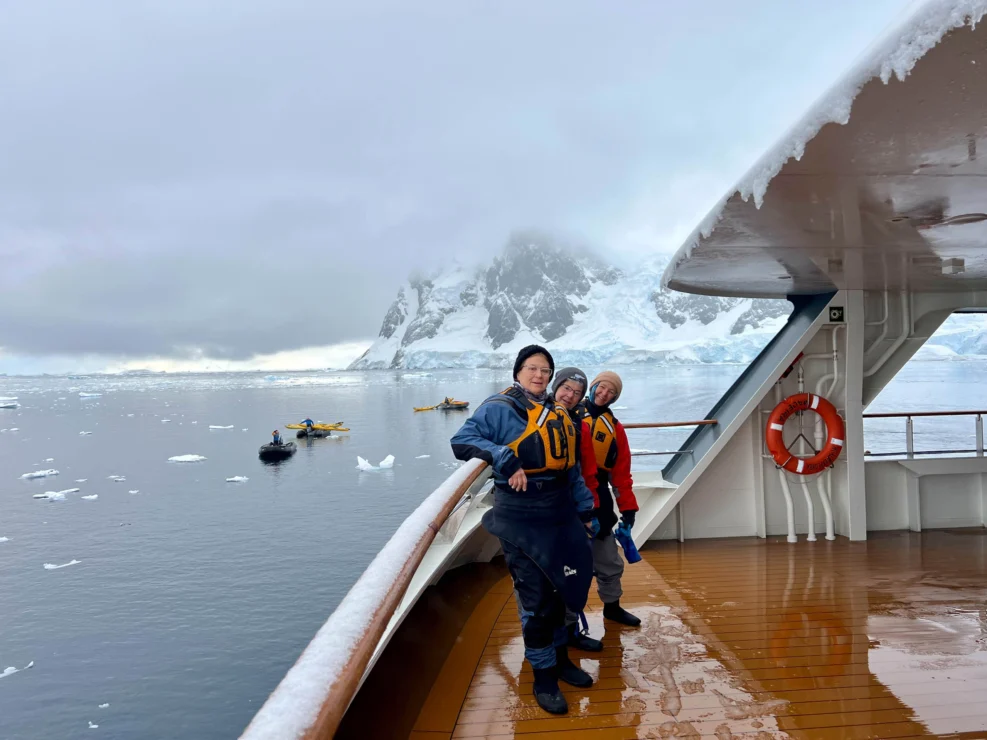 Three people in winter gear stand on a ship deck overlooking an icy body of water with snow-covered mountains in the background.