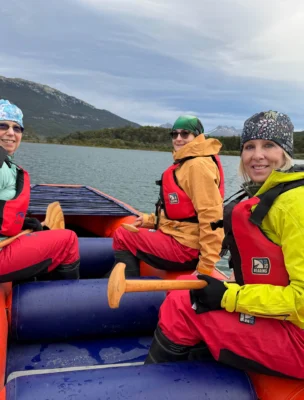 Three people in colorful jackets and life vests paddle a raft on a lake with mountains in the background.