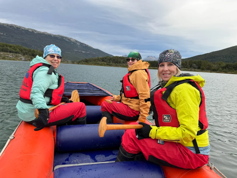 Three people in colorful jackets and life vests paddle a raft on a lake with mountains in the background.