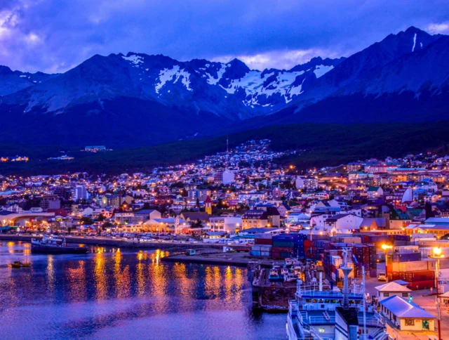 A vibrant coastal city at dusk with illuminated buildings and mountains in the background. The waterfront reflects the city lights, and the sky is a mix of blue and purple hues.