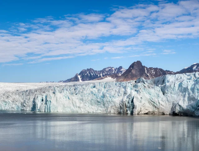 A panoramic view of a glacier meeting a calm body of water with a background of rugged mountains under a partly cloudy blue sky.