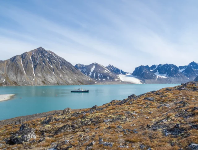 Mountainous landscape with a ship in a bay, surrounded by snowy peaks and partly cloudy skies. Foreground features rocky terrain with sparse vegetation.