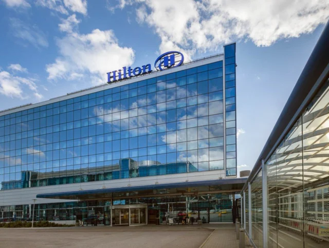 View of a Hilton hotel building with a glass facade reflecting the sky, featuring the Hilton logo on top under a partly cloudy sky.