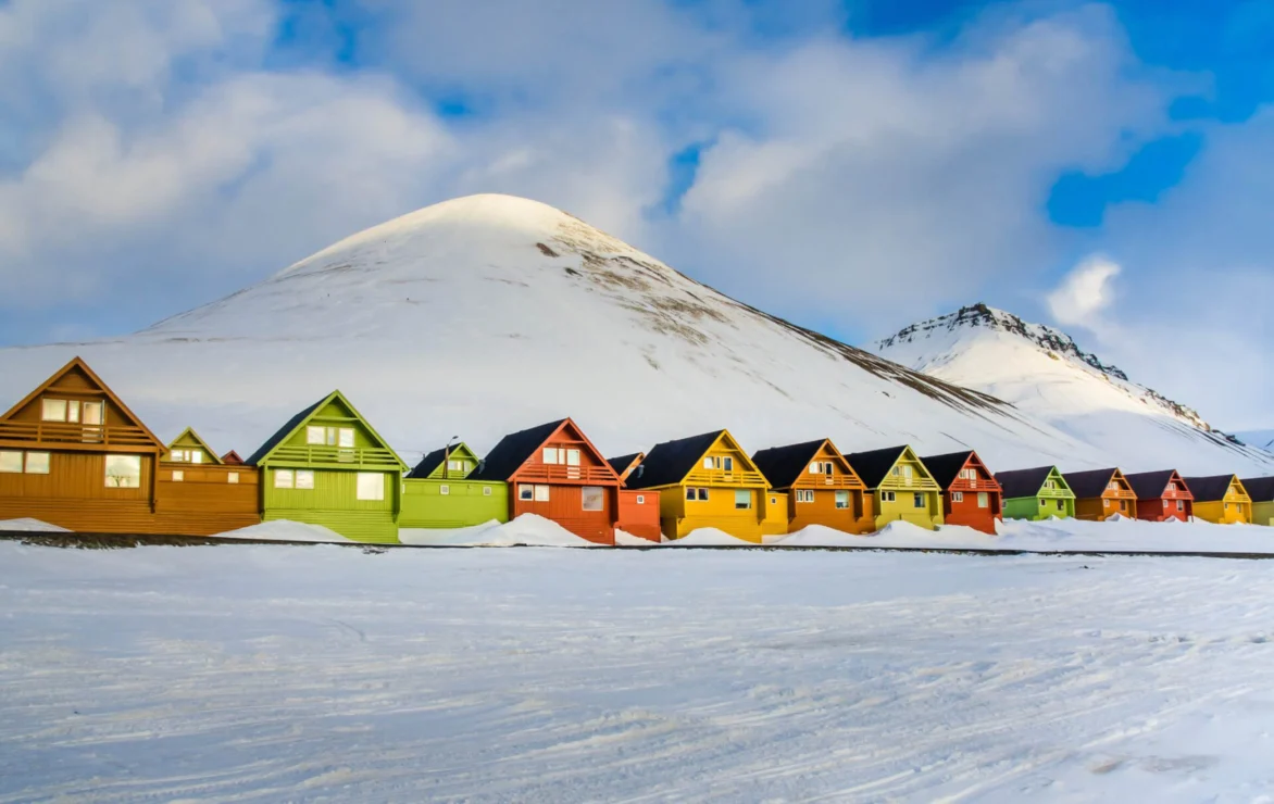 A row of colorful, triangular-roofed houses stand against a snowy mountain backdrop under a cloudy sky.