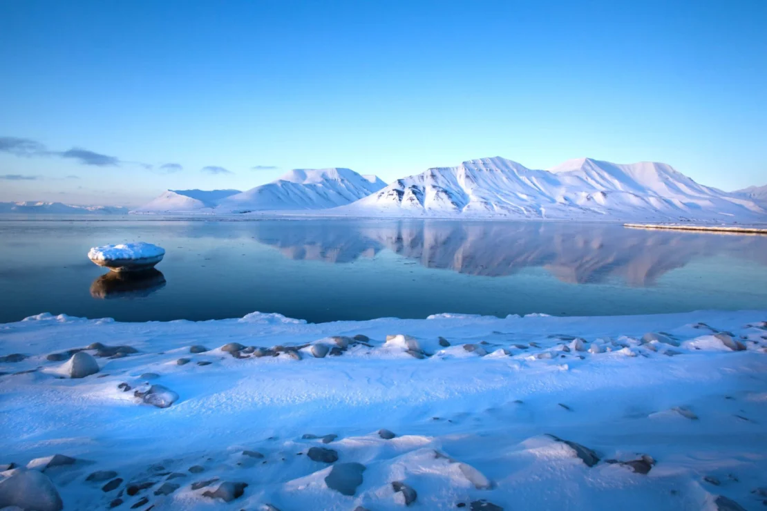 Snow-covered landscape with a calm body of water reflecting snowy mountains under a clear blue sky. Rocks and ice are visible along the shoreline.