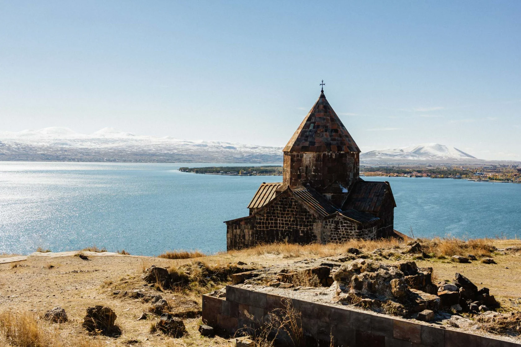 A historic stone church stands on a grassy hill overlooking a large blue lake, with distant snow-capped mountains under a clear sky.