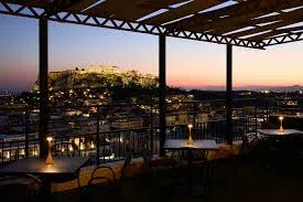 Rooftop restaurant terrace at dusk with tables and candles, overlooking a cityscape and a lit ancient hilltop structure in the background.