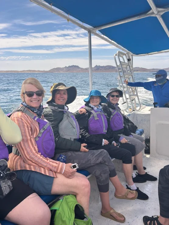 Four people wearing life jackets smile on a boat under a canopy, with ocean and mountains in the background.