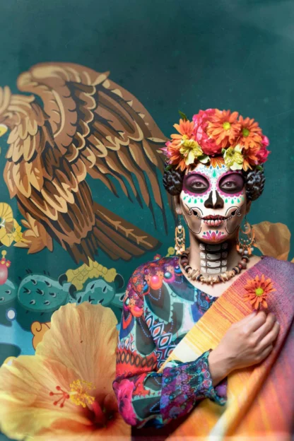 Person with Day of the Dead face paint, wearing a colorful floral headdress and attire, stands against a vibrant backdrop of an eagle and floral designs.