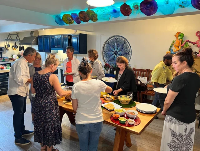 People are gathered around a dining table with various dishes, preparing food in a kitchen with colorful decor.