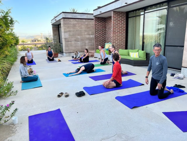 A group of people doing yoga on mats outside a modern building, with a few seated and others stretching.