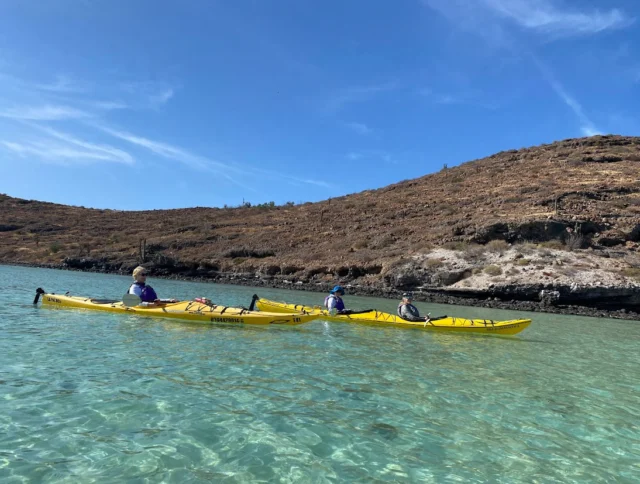 Three people are kayaking in clear, shallow water with a rocky hillside in the background under a blue sky.