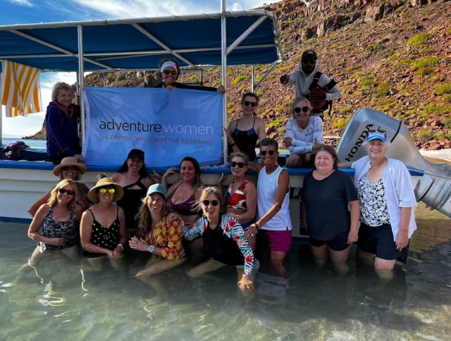 A group of women pose in front of a boat with "Adventure Women" signage, standing in shallow water near a rocky shore.