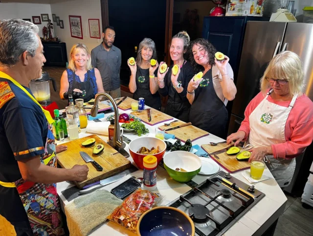 Group of people in a kitchen holding halved avocados, surrounded by ingredients on the counter, smiling and preparing food together.