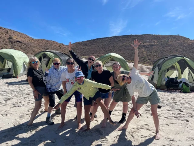 A group of eight people posing energetically on a sandy beach with green tents and hills in the background under a clear blue sky.