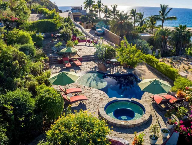 Aerial view of a landscaped backyard with a pool, hot tub, lounge chairs, and green umbrellas. The scene is surrounded by lush greenery and overlooks the ocean in the background.