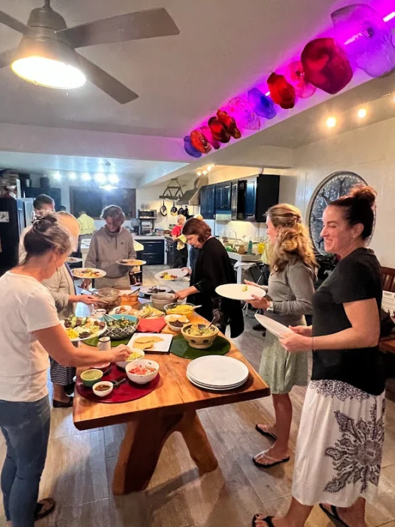 A group of people serving themselves food from a buffet-style table in a kitchen with colorful caps on a shelf above.