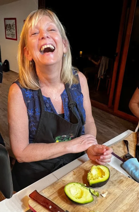 Smiling woman wearing an apron stands at a table with a halved avocado on a cutting board.