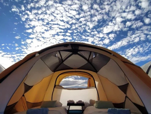 Interior of a spacious tent with two beds and a small table under a sky with scattered clouds.