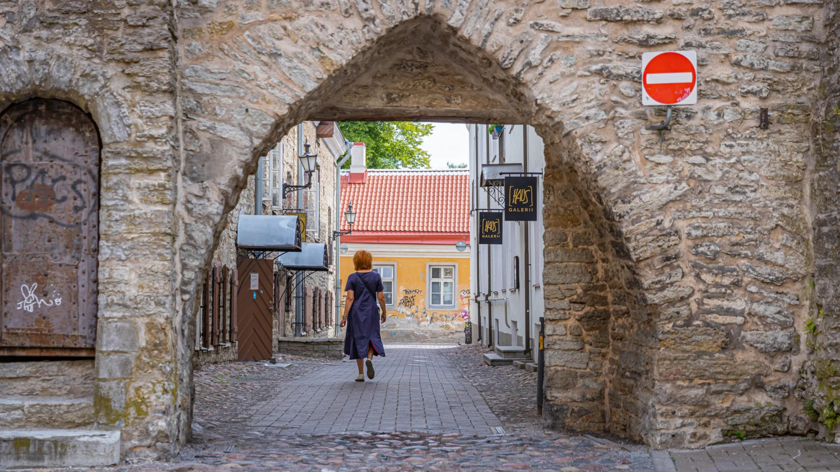 Person walks through a stone archway on a cobblestone street in a historic area, with signs and old buildings in the background.