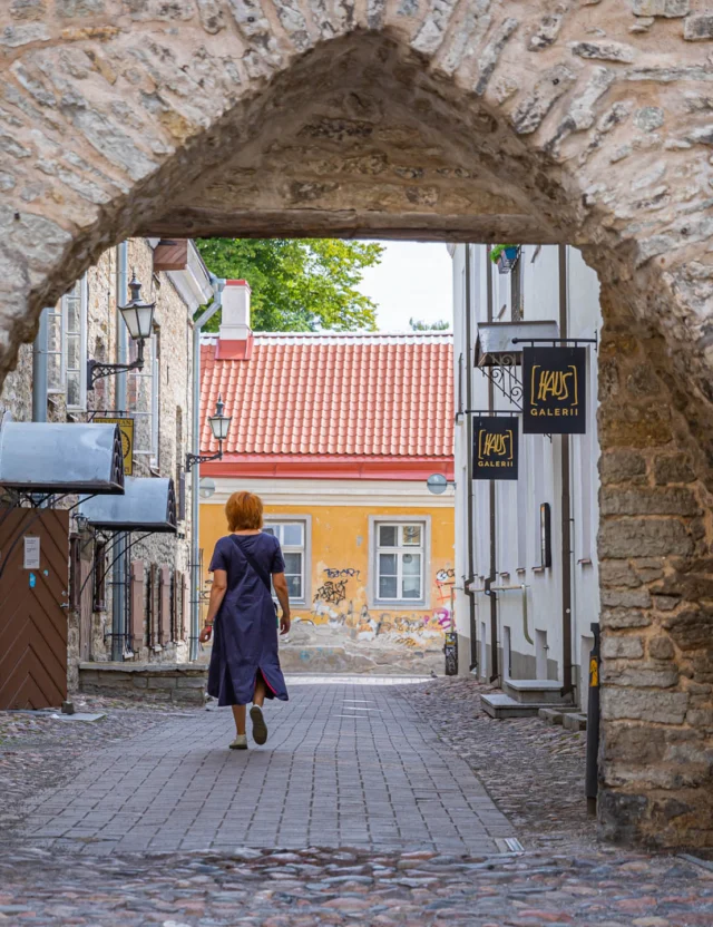 Person walks through a stone archway on a cobblestone street in a historic area, with signs and old buildings in the background.