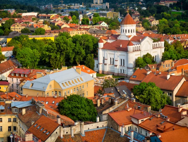 Aerial view of a European town with colorful buildings, red roofs, and a large white church surrounded by greenery.