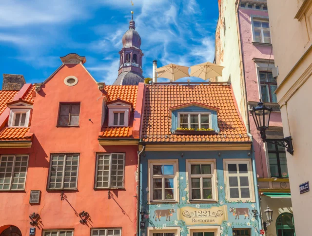 Colorful historic buildings with orange tiled roofs under a blue sky, featuring a church spire in the background and a restaurant sign on a pastel facade.