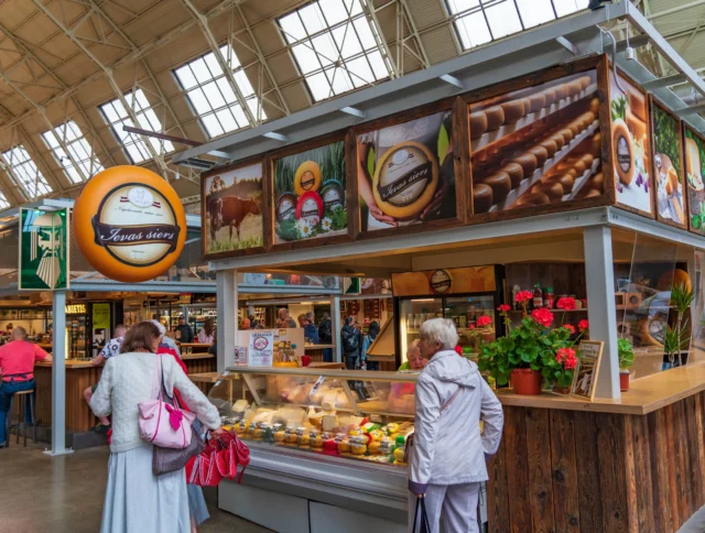 Two people stand in front of a cheese display at an indoor market stall with large overhead signs and potted flowers.