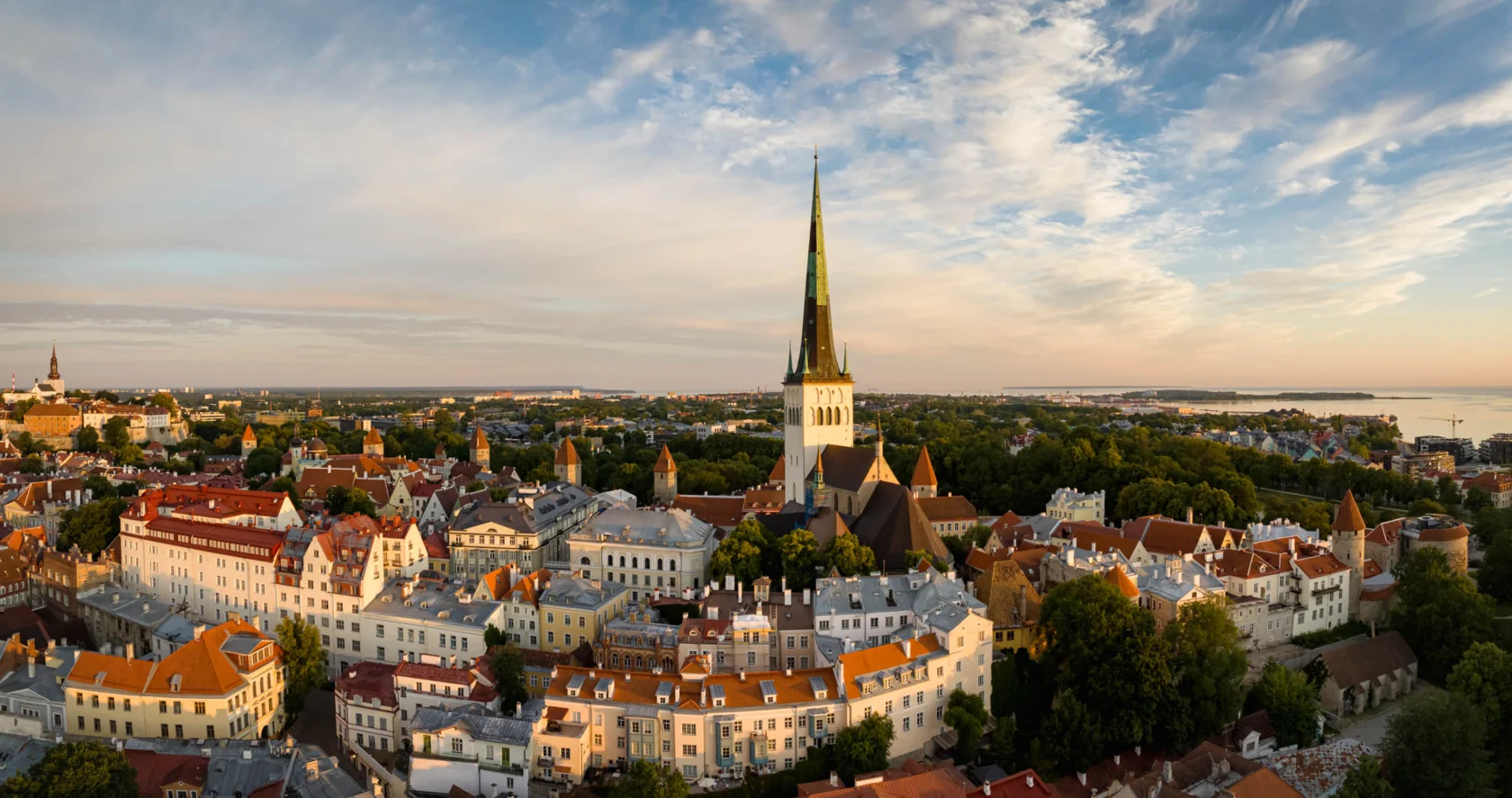 Aerial view of a historic cityscape with a tall church spire, surrounded by red-roofed buildings and greenery under a partly cloudy sky.