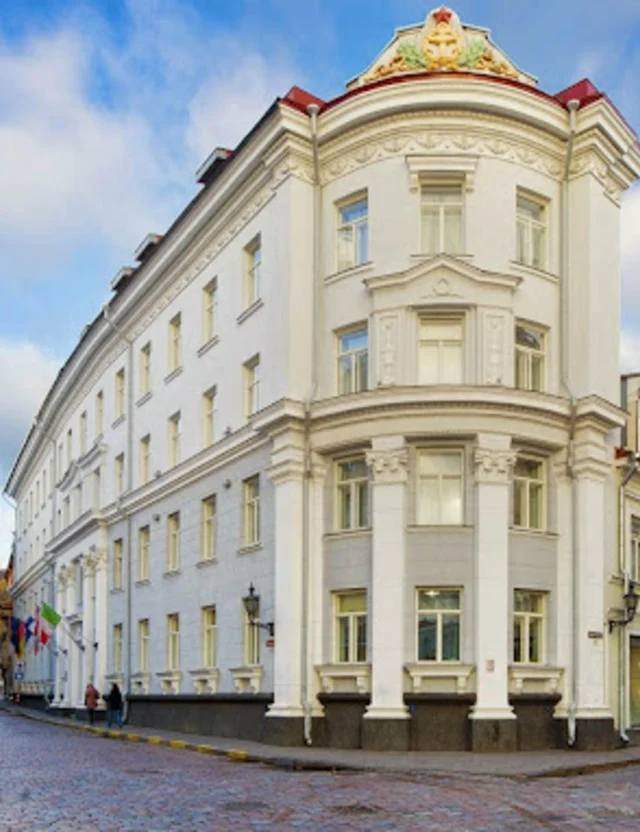 A large white historic building stands at a street corner with cobblestone roads, under a partly cloudy sky.