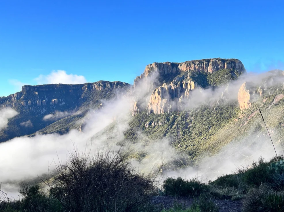 Mountain landscape with cliffs and green vegetation partially covered by clouds, under a clear blue sky.