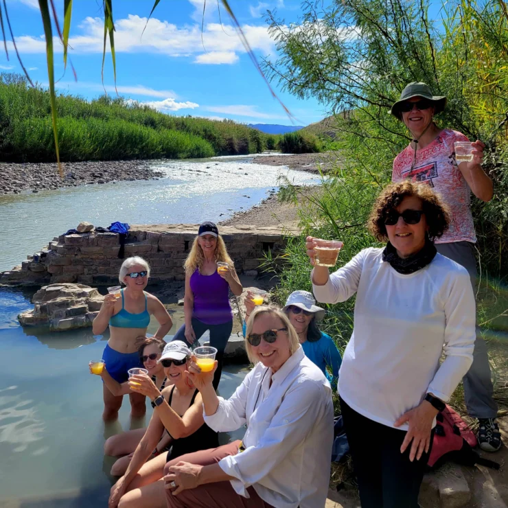 A group of people enjoying drinks by a riverside, some standing and others sitting in the water, surrounded by greenery under a clear sky.