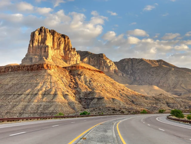 A winding road leading towards a rocky mountain peak under a partly cloudy sky.