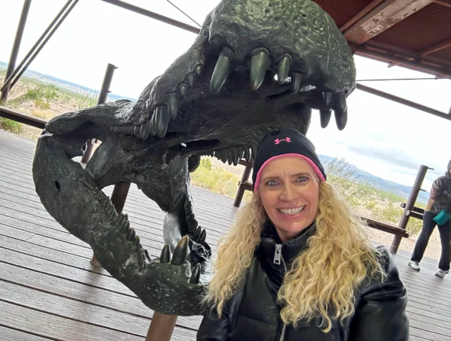 A woman with curly blonde hair poses smiling next to a large dinosaur skull exhibit on a wooden deck.